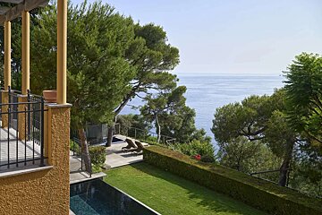 A luxurious Mediterranean villa garden with a pool, lush lawn, pine trees, and a breathtaking view of the sparkling blue sea.