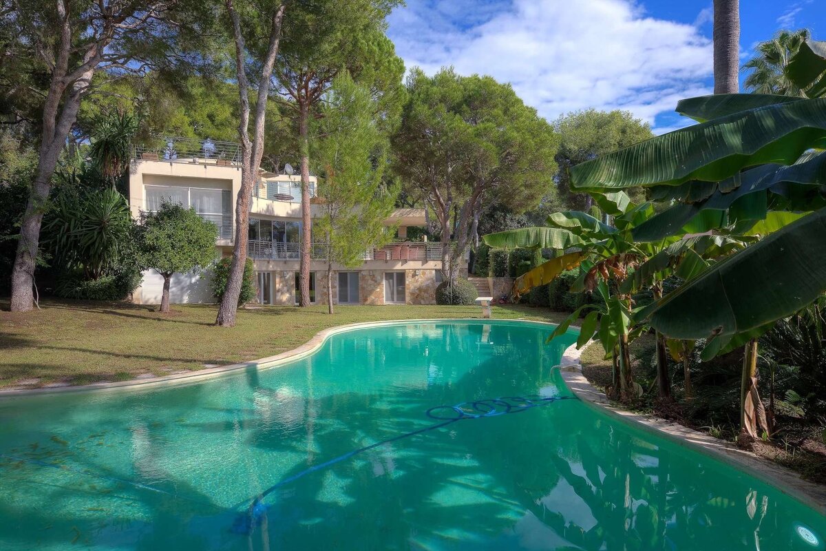 A large villa with a curved turquoise pool, surrounded by lush trees and green lawn under a partly cloudy sky. A pool cleaner is visible.