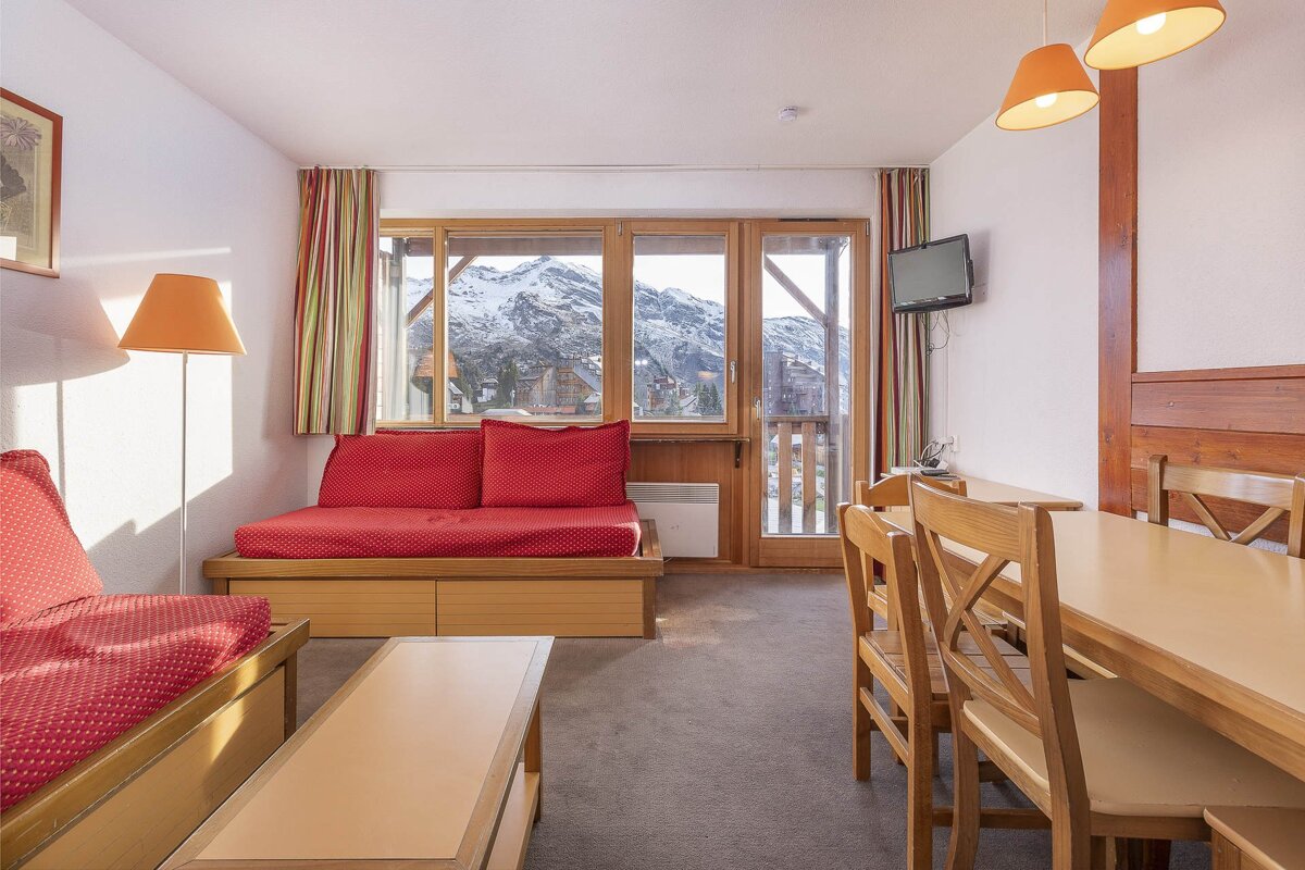 A cozy mountain apartment living/dining area with red sofa beds, wooden furniture, and a large window overlooking snow-capped peaks and a village.