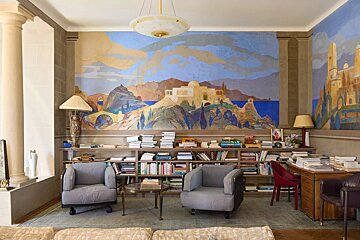 A cozy study featuring low bookshelves, comfortable grey armchairs, and a large vibrant landscape mural on the wall.