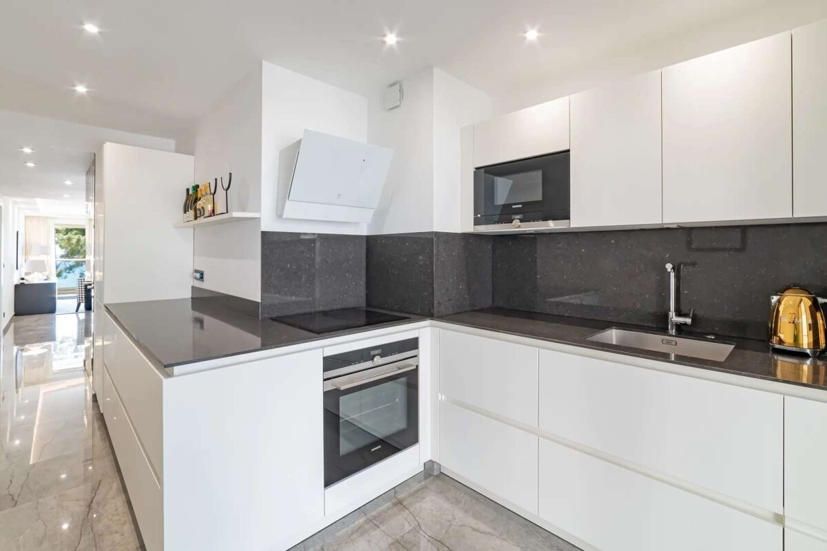 A modern, bright white kitchen features dark countertops, built-in appliances like an oven and microwave, and a golden kettle. It has an open-plan layout.
