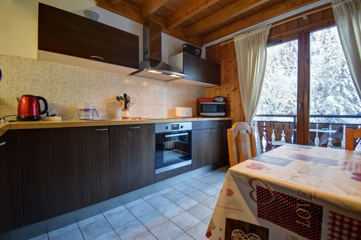 Cozy chalet kitchen and dining area with dark cabinets, appliances, and a table. A large window overlooks a snowy forest and balcony.