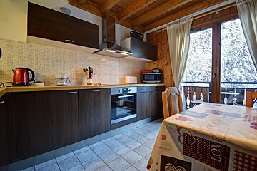 A chalet-style kitchen and dining area with modern appliances, a wooden ceiling, and a balcony view of snow-covered trees.