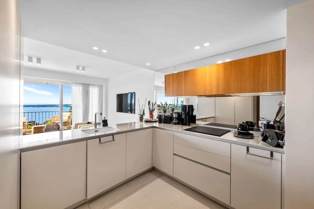 Sleek white and wood kitchen with a mirrored backsplash. It opens to a bright balcony featuring a stunning sea or lake view.