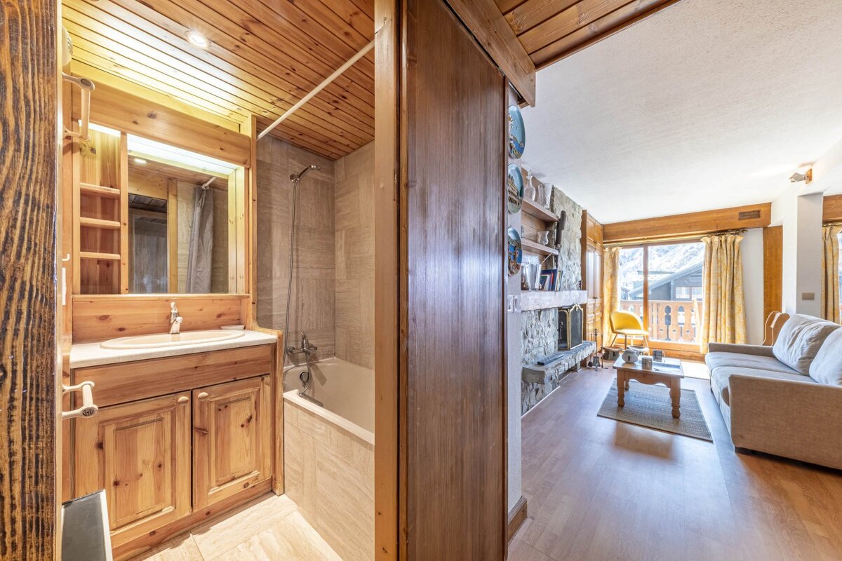 A rustic interior featuring a wood-paneled bathroom with a tub and vanity on the left, and a cozy living room with a stone fireplace and sofa on the right.
