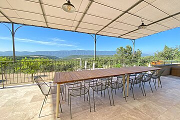 A spacious outdoor patio with a long dining table and chairs under a canopy, offering a panoramic view of mountains and a lush valley.