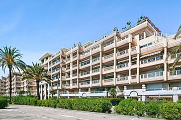 A large, multi-story pink building with numerous balconies and rooftop terraces, flanked by palm trees and green hedges under a clear blue sky.