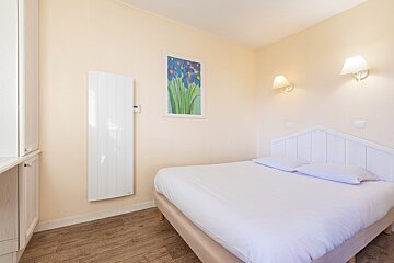 A bright bedroom with a white bed, wooden headboard, wall lamps, floral painting, wood floor, and a white heater.