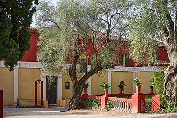 A striking red and yellow building, adorned with classic text, sits behind lush olive trees. A red balustrade and potted plants enhance the scenic facade.