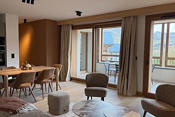 Modern chalet interior with a dining area, cozy armchairs, and large glass doors opening to a scenic balcony.