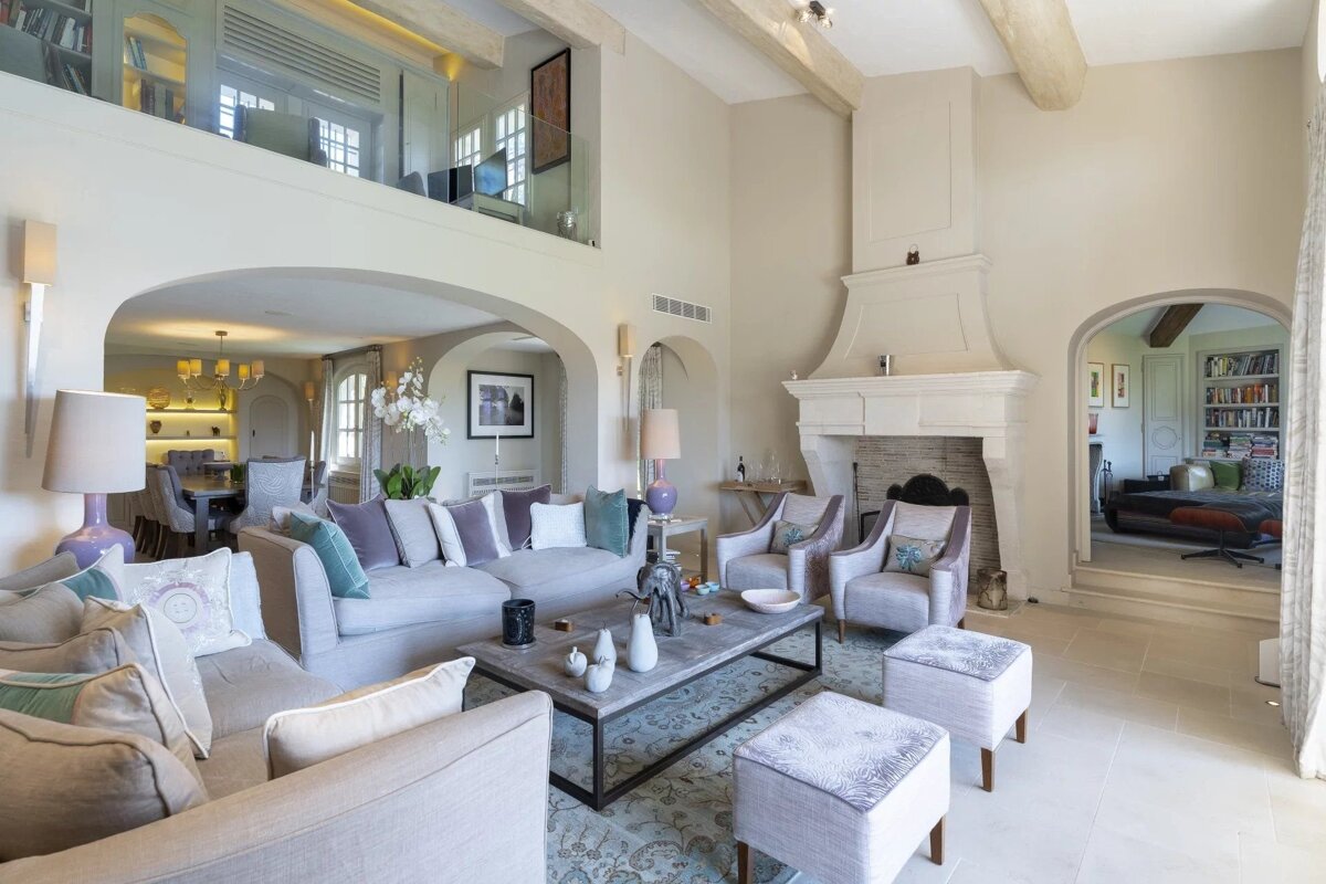 A spacious, elegant living room with neutral sofas, a stone fireplace, high beamed ceilings, and a mezzanine balcony.