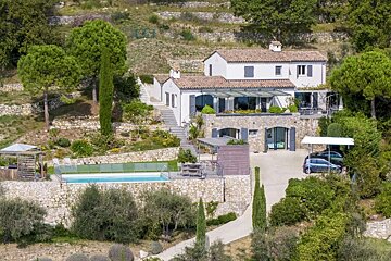 A white house on a terraced hillside with a swimming pool, lush greenery, and cars parked under a carport.