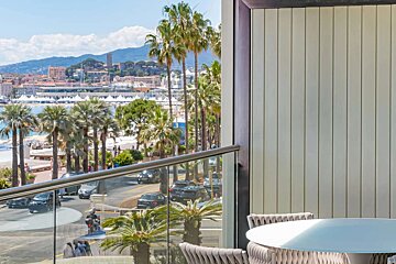 View from a balcony with table and chairs, overlooking a palm-lined coastal town with a harbor, cars on a road, and buildings on a distant hillside.