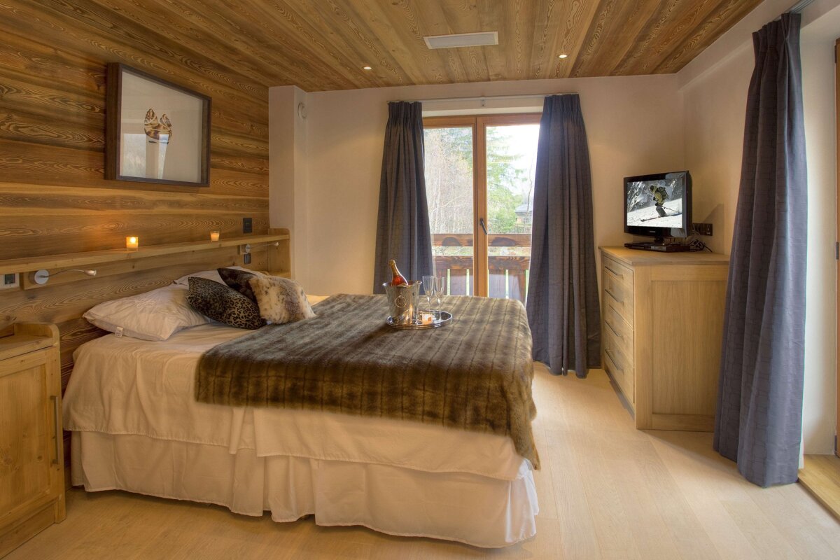 Cozy ski chalet bedroom with wood paneling, a large bed, champagne on a tray, balcony view, and a TV showing skiing.