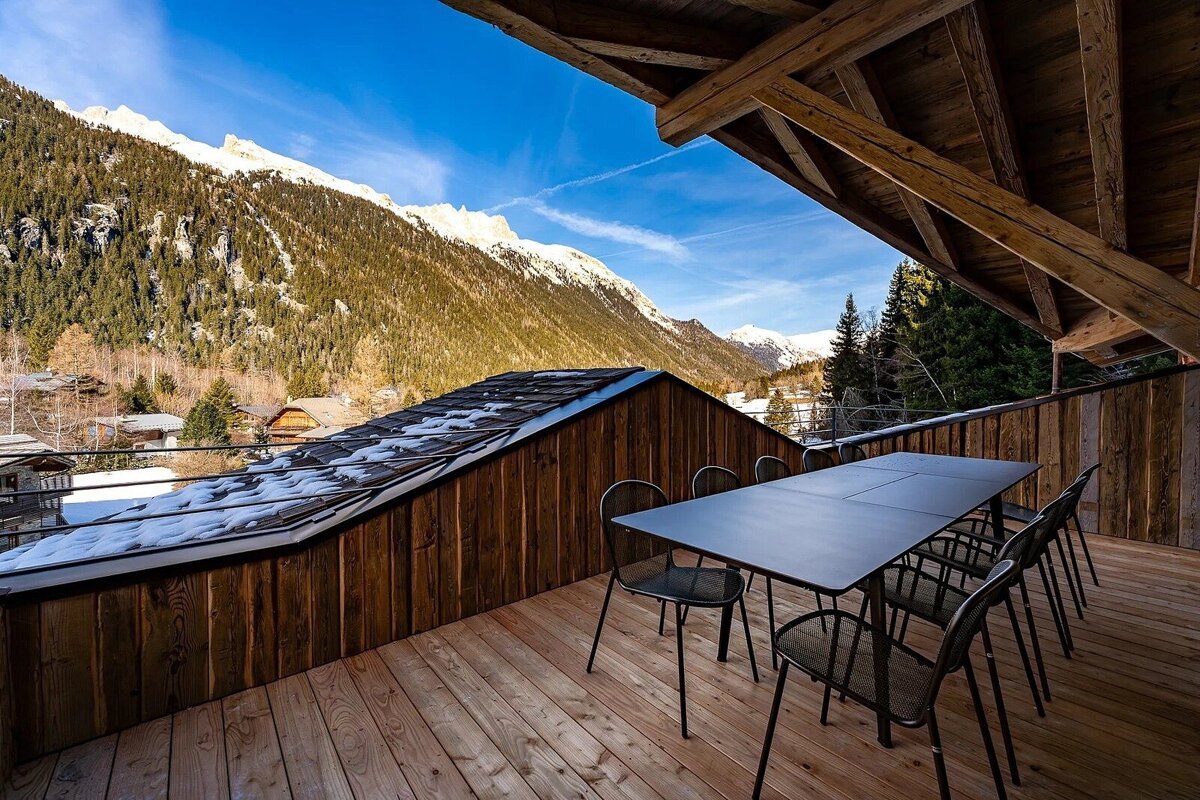 Wooden chalet balcony with a dining table overlooking stunning snow-capped mountains and evergreen forests.