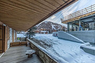 A snowy mountain ski resort village seen from a rustic wooden deck with an overhead wooden roof, featuring distant slopes and a modern building under construction.