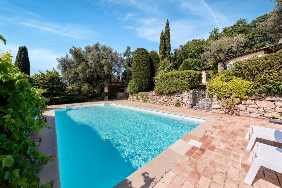 A pristine blue swimming pool with terracotta tiles, surrounded by lush green trees, shrubs, and stone walls, under a clear, sunny sky.