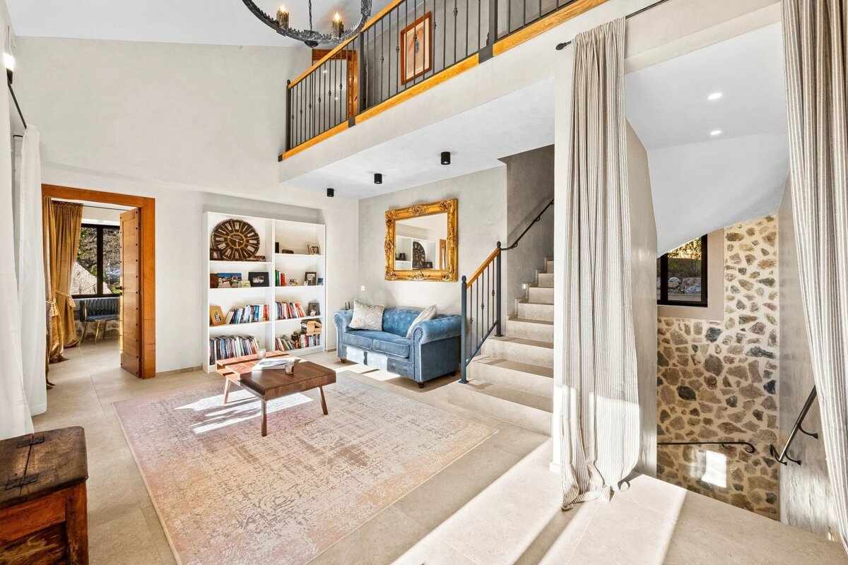 A spacious, sunlit living room with high ceilings, a mezzanine balcony, blue sofa, bookshelves, and stone-accented stairs.