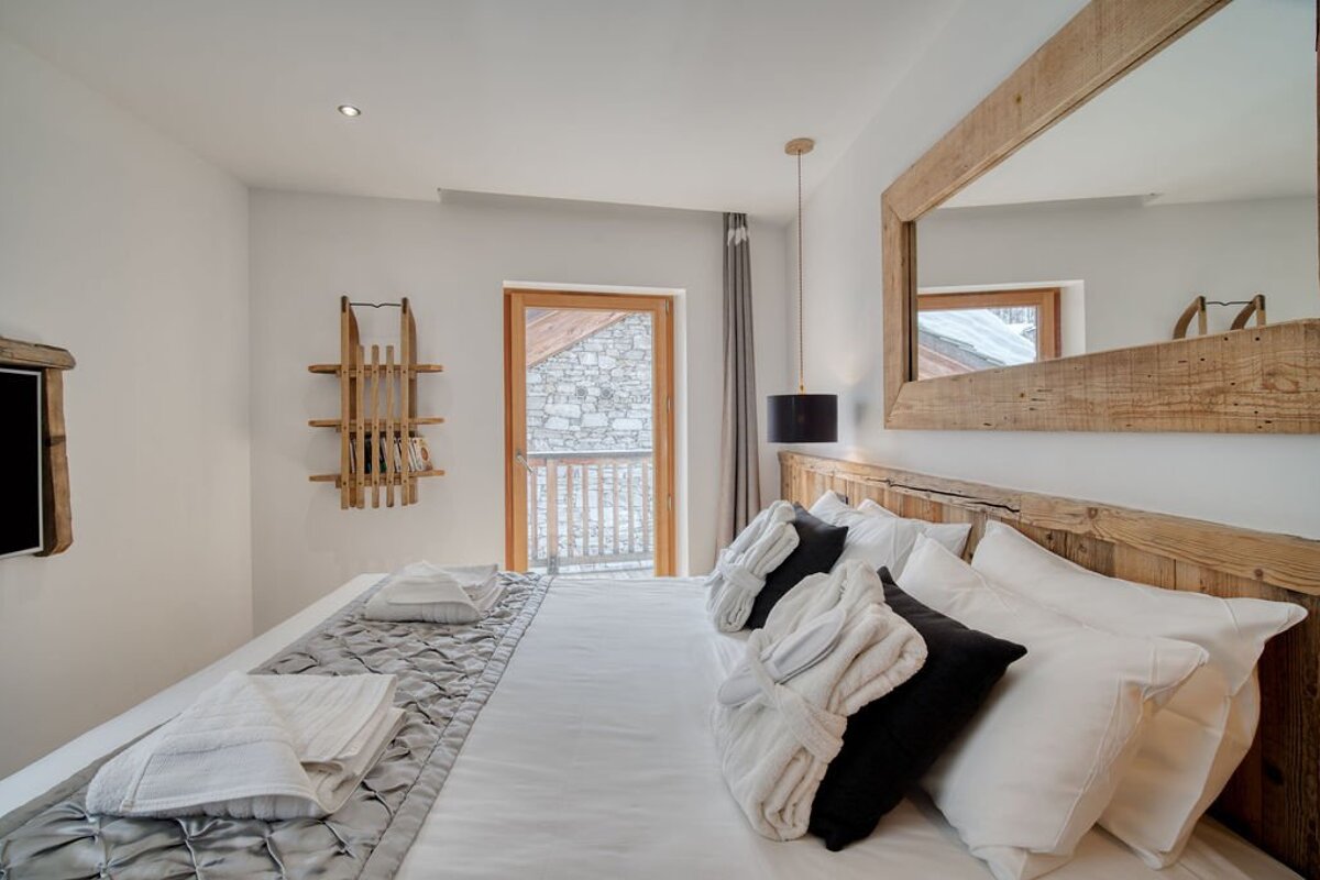 Cozy chalet bedroom featuring a bed with folded robes, a rustic wooden headboard, a matching mirror, and a door leading to a balcony.