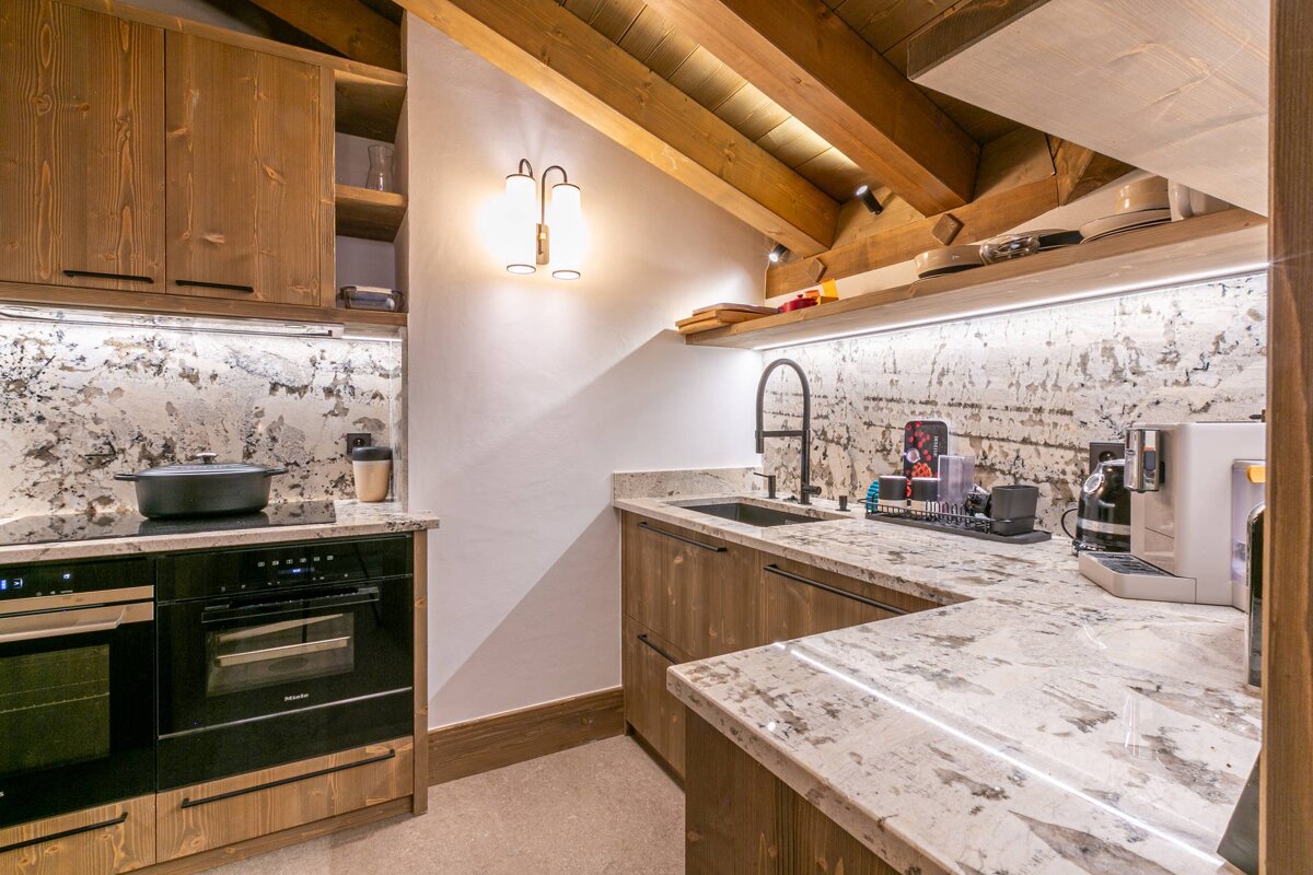 Cozy attic kitchen with warm wood cabinets, granite countertops & backsplash, exposed beams, and modern black appliances, illuminated by accent lighting.