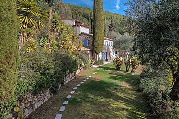 A rustic stone house with blue shutters sits amidst a vibrant, sunlit garden with a stepping stone path, tall trees, cacti, and green hills.