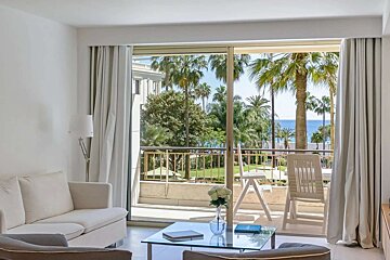 White living room with a large sliding door opening to a balcony overlooking palm trees, a green garden, and the blue ocean.