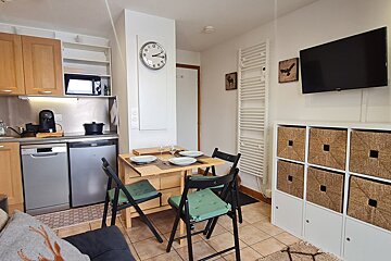 A compact studio apartment featuring a modern kitchenette, a wooden dining table for four, a wall-mounted TV, and a white storage unit with woven baskets.