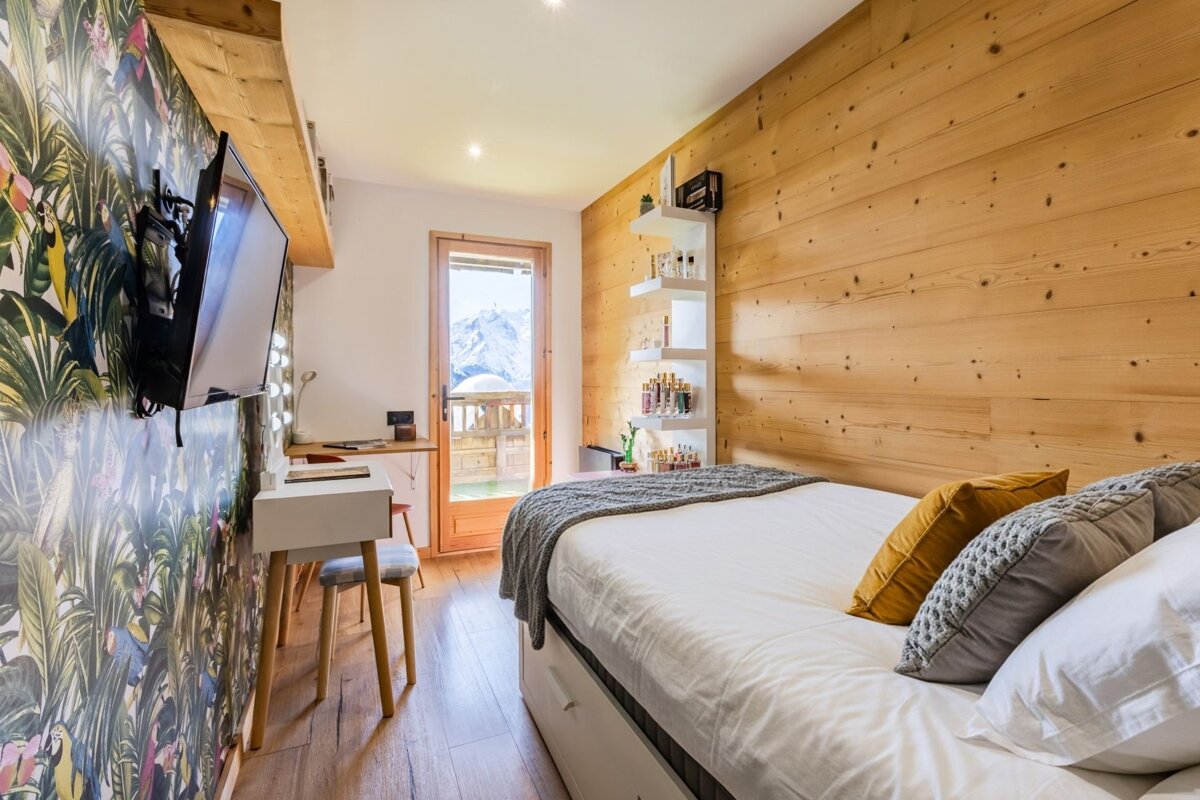 Cozy chalet bedroom with wood paneling, tropical wallpaper, a small vanity, and a balcony showing snowy mountain views.