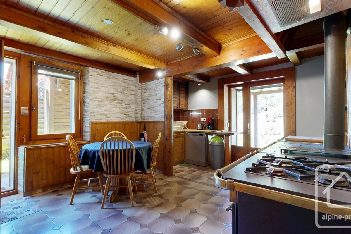 A rustic kitchen-diner with rich wooden beams, ceiling, and wainscoting. Features a tiled floor, round dining table, and a large, traditional stove.
