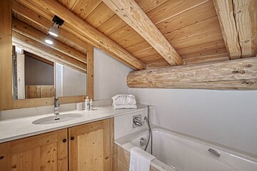 Attic bathroom with slanted wooden ceiling & exposed beams over a vanity, mirror, and white bathtub. Rustic-modern design.