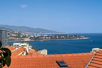 View over blue sea and green coast with distant mountains from red tiled rooftops. Buildings dot the landscape under a clear sky.