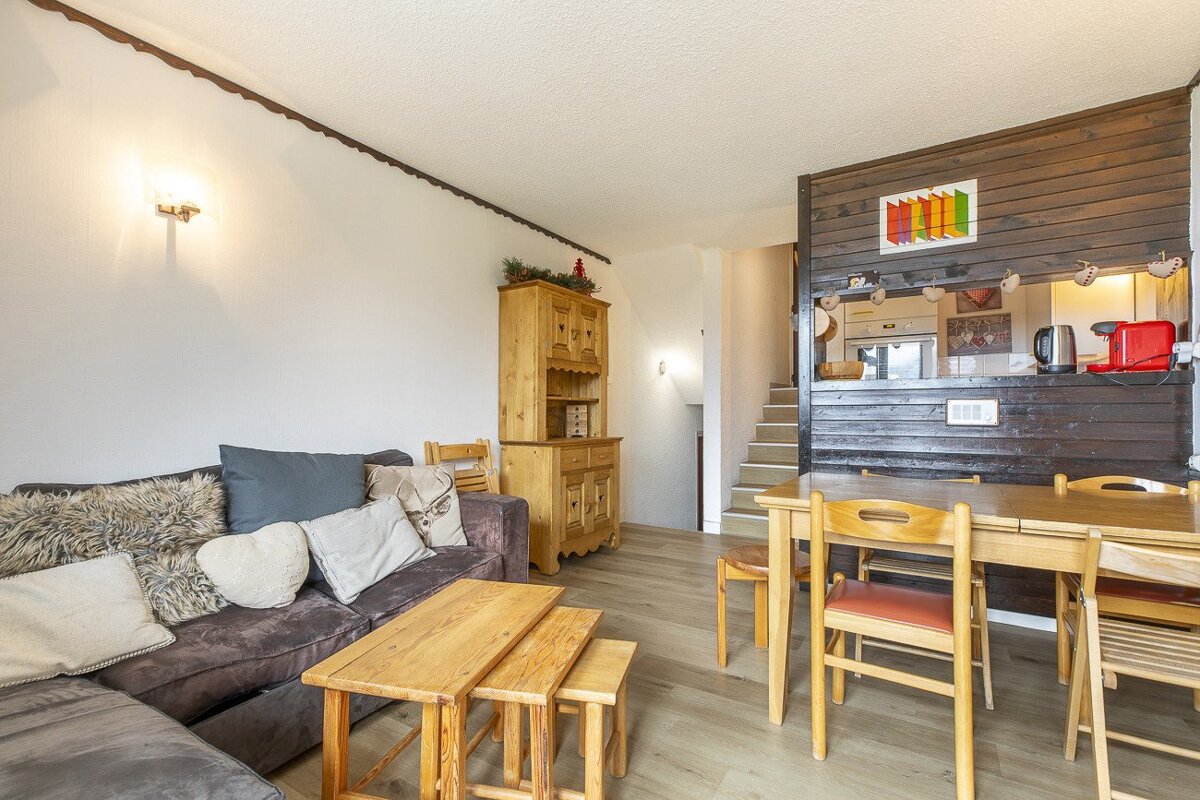 An inviting chalet-style living and dining room with a sofa, wooden tables, a large hutch, and a dining set. A wood-paneled kitchen bar and stairs are visible.