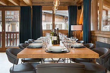 A wooden dining table set with plates, glasses, and wine in a cozy chalet with modern grey chairs and dark blue curtains.