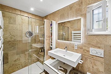 Modern bathroom with beige stone tiles, a glass-enclosed shower, a floating white sink, and a backlit mirror.