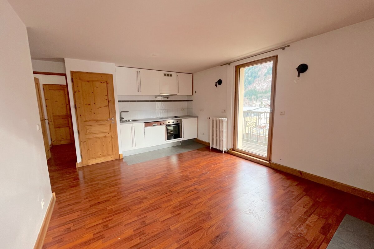 Bright, empty apartment with a compact white kitchen, polished wooden floor, and a balcony offering a scenic mountain view.