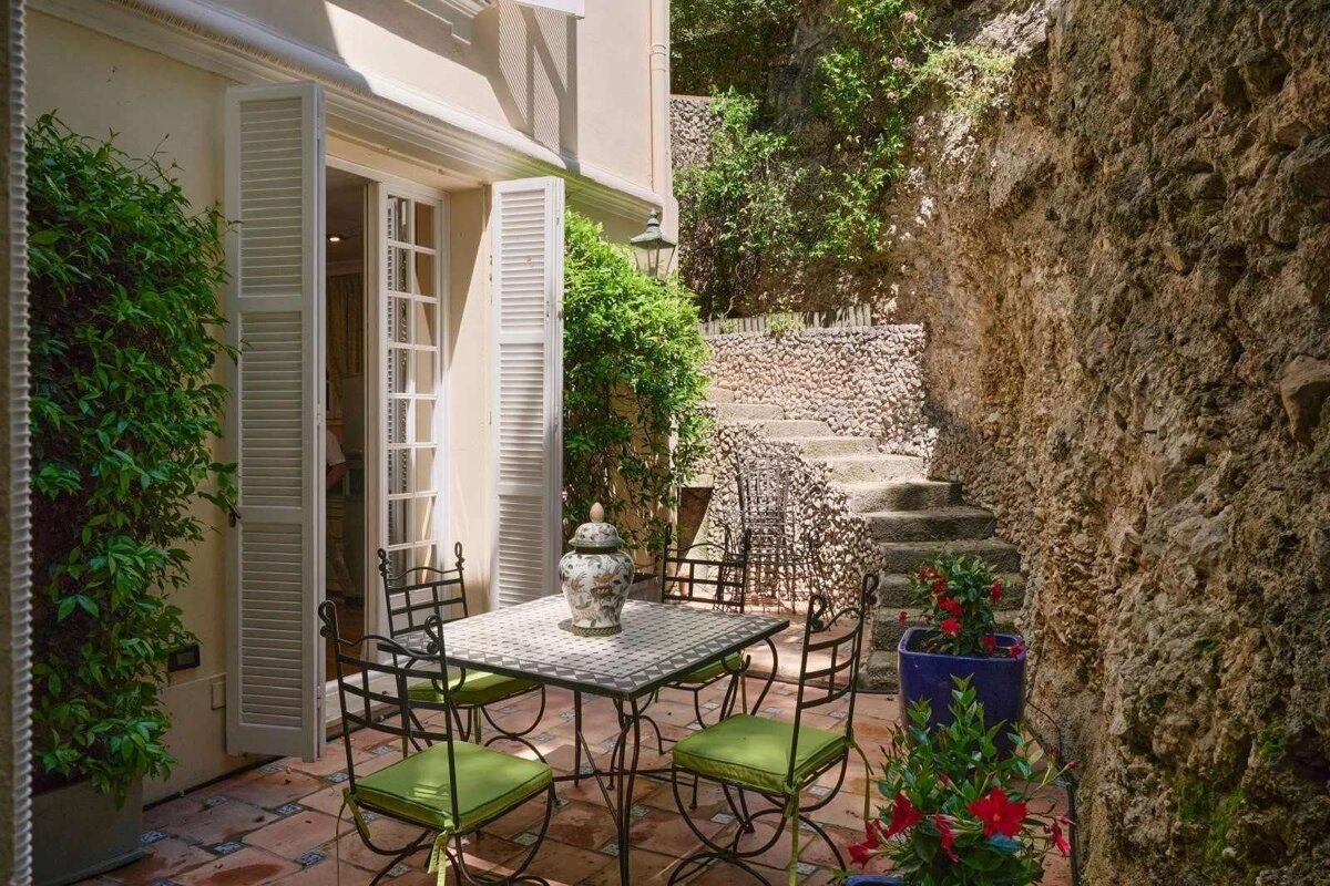 A sunny Mediterranean patio with a wrought iron table and chairs, terracotta tiles, open shutters, and stone steps leading up a lush, rocky hillside.