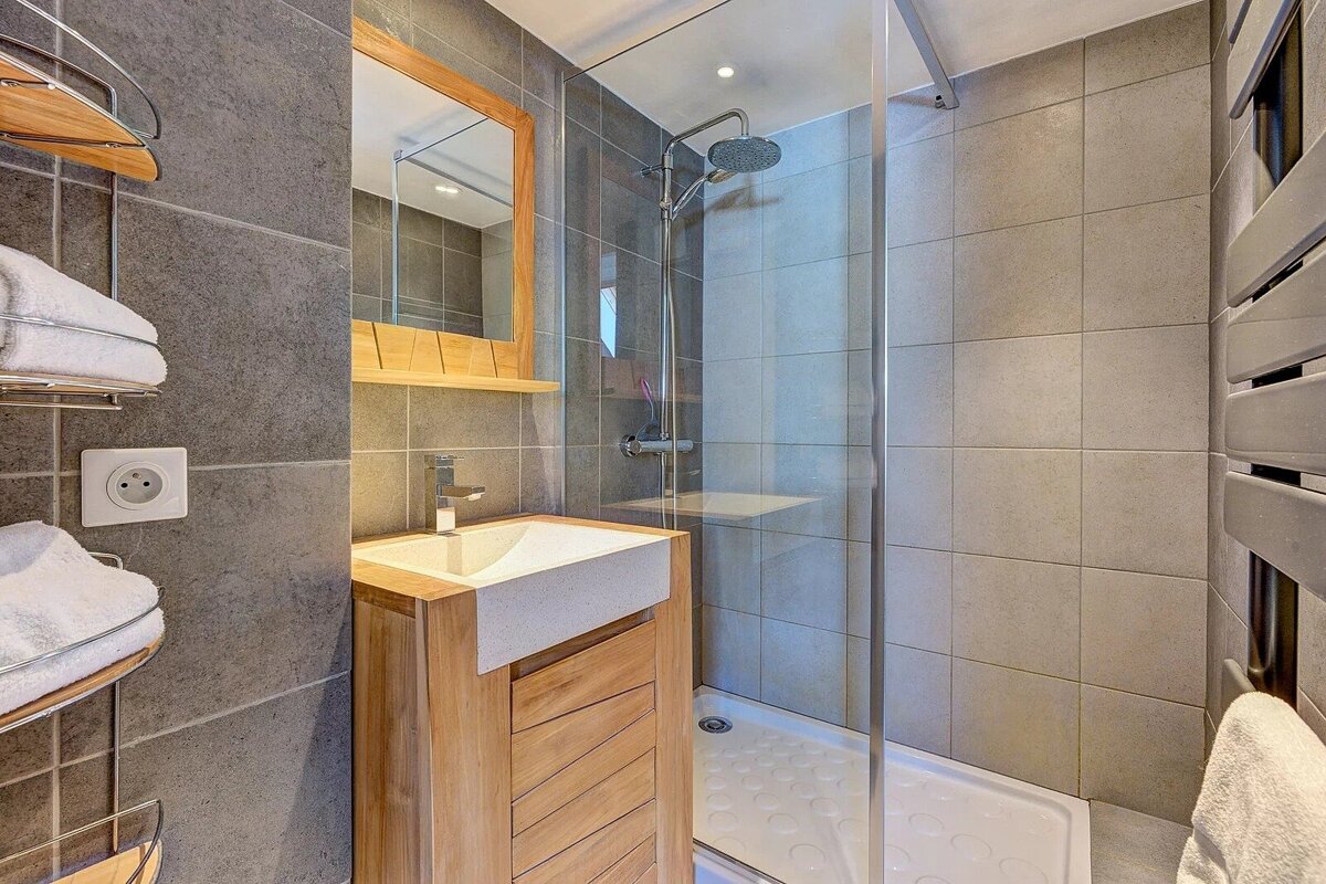 Modern bathroom featuring grey tiled walls, a wooden vanity with sink, a framed mirror, and a glass-enclosed shower.