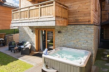 A cozy wooden chalet deck featuring a steaming hot tub, comfortable outdoor seating, and a stone facade.