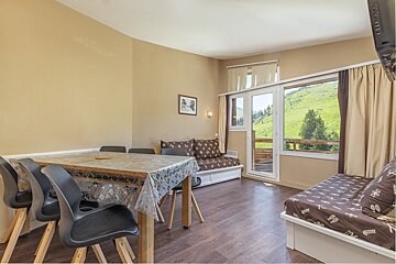 A cozy apartment living room with a dining table, two beds, and large windows offering a beautiful view of lush green mountains.
