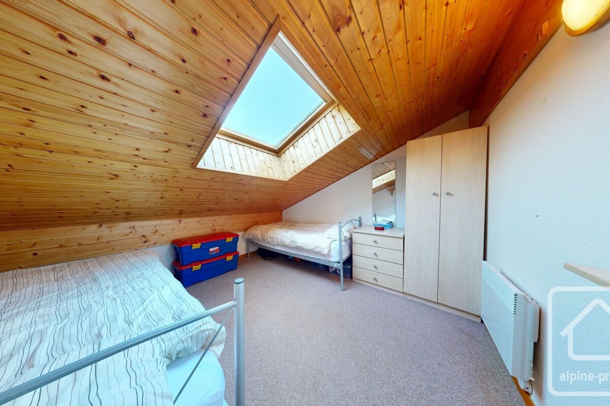 An attic bedroom featuring a sloped wooden plank ceiling, a bright skylight, two single beds, and light-toned wardrobe and dresser.