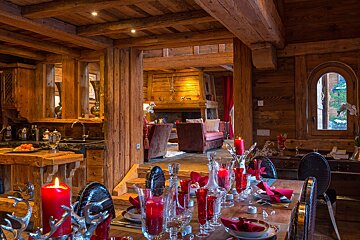 Cozy timber chalet interior: red-themed dining table in the foreground, with a warm living area and fireplace in the background.