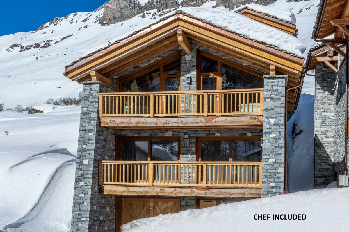 A stone and wood ski chalet with balconies against a snowy mountain backdrop. 