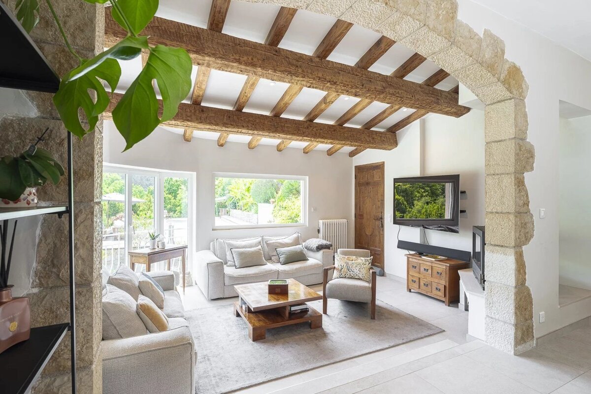 Bright rustic living room with exposed wood ceiling beams, cozy grey sofas, and a stone archway.