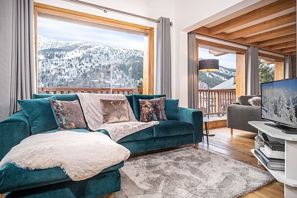 Cozy living room with a teal sofa, large windows overlooking a snowy mountain landscape, and a balcony view.