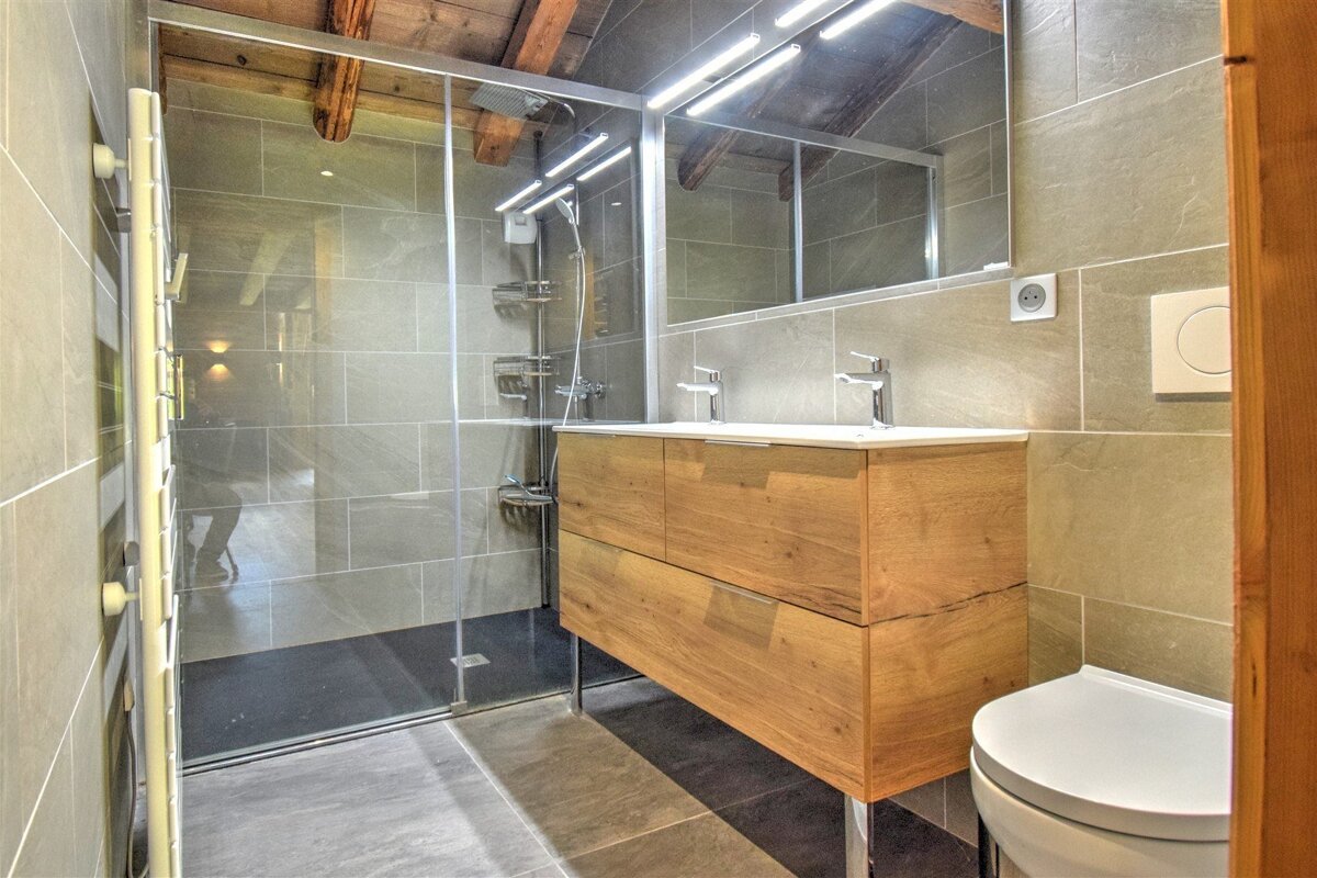 A modern bathroom featuring gray tiled walls and floor, a glass shower enclosure, a double wooden vanity with sinks, and a toilet. Exposed wooden beams are visible overhead.