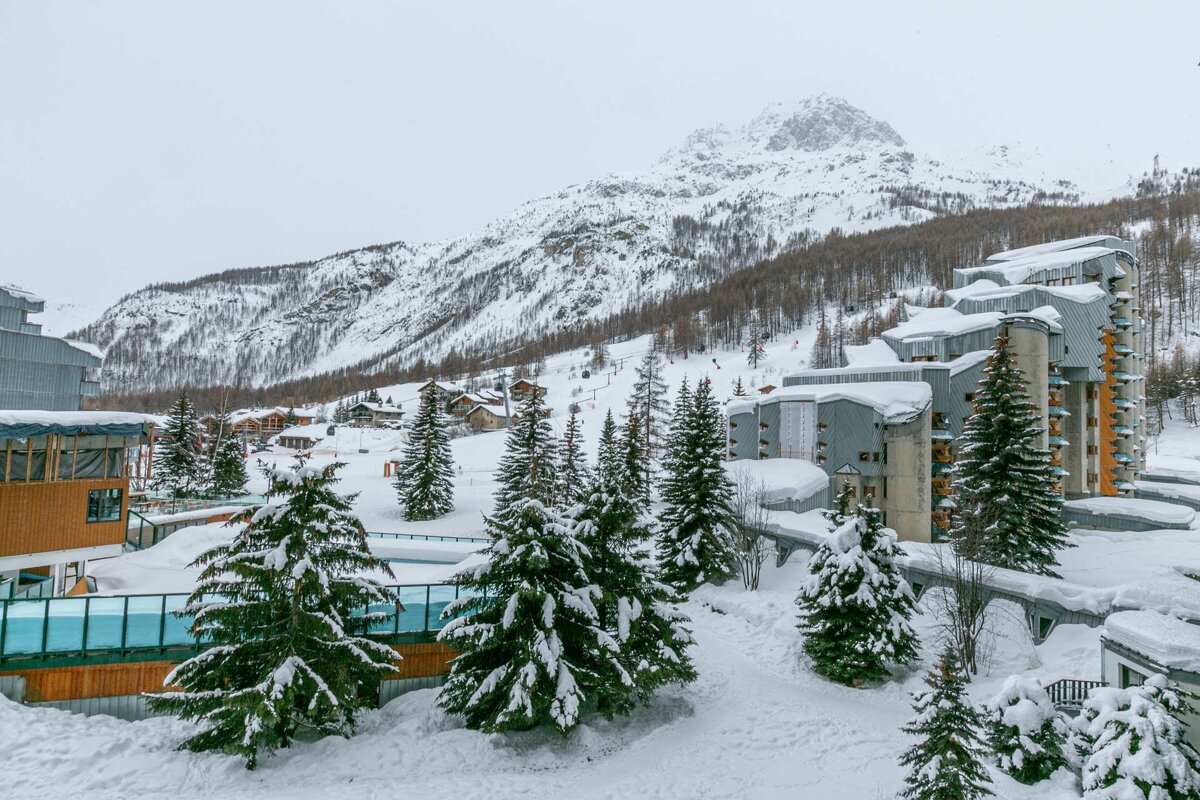 A snowy mountain ski resort features modern buildings, evergreen trees, and a majestic peak under a cloudy winter sky.