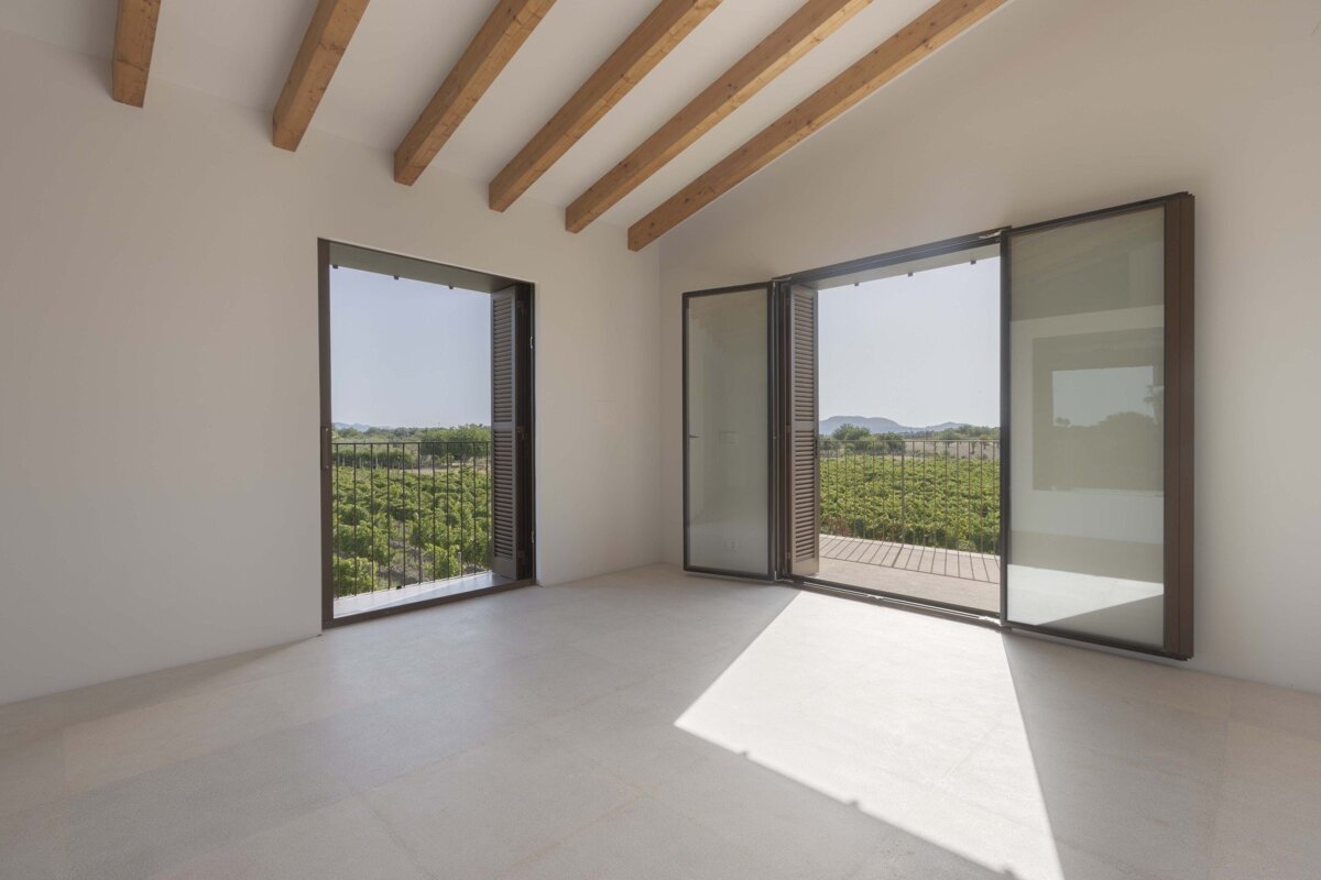 Minimalist white room with wooden beams and two large doorways opening to a lush vineyard and distant mountains under sunlight.