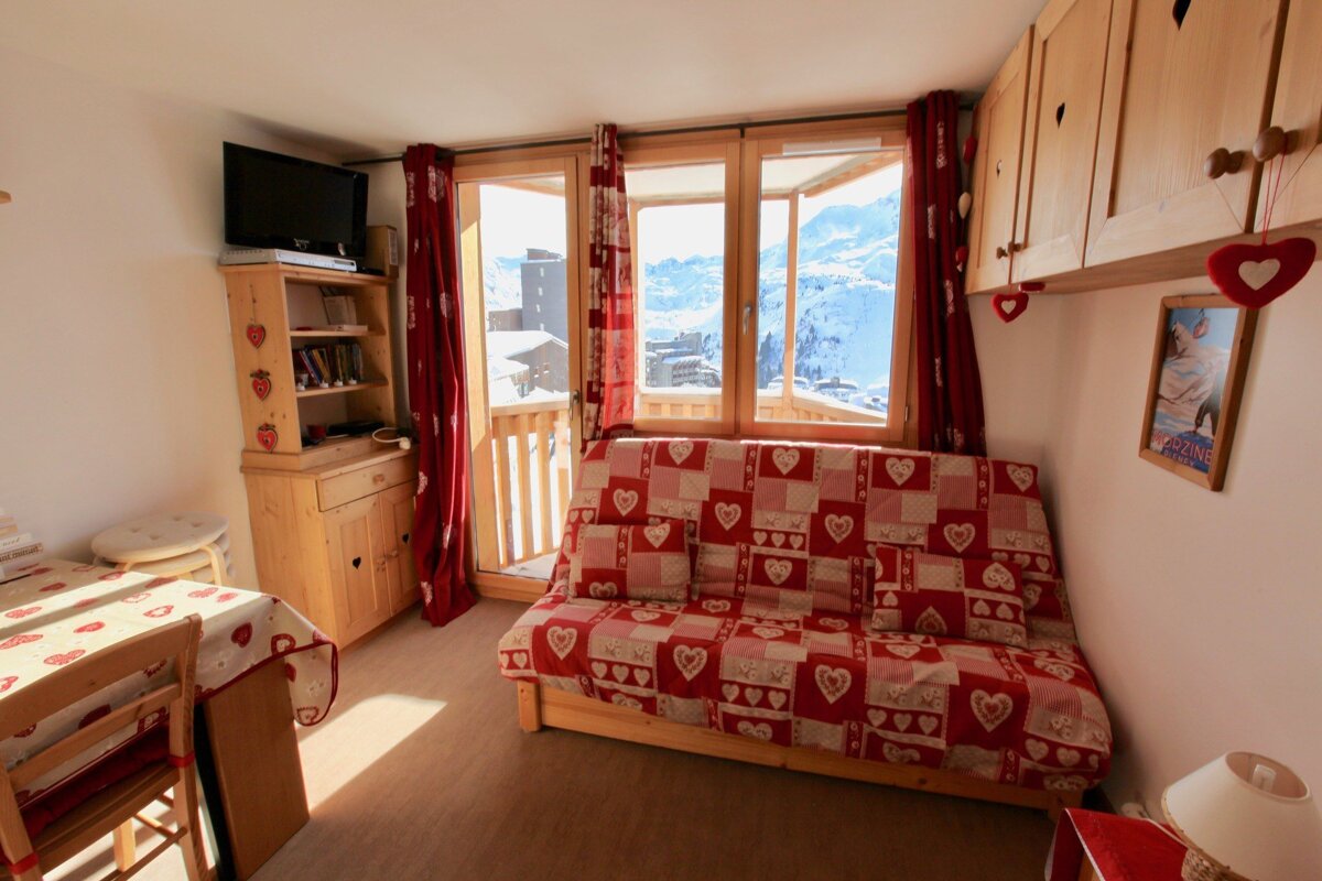 Cozy ski apartment with a red heart-patterned sofa, wooden furniture, and a balcony offering a stunning view of snow-covered mountains and a ski resort.