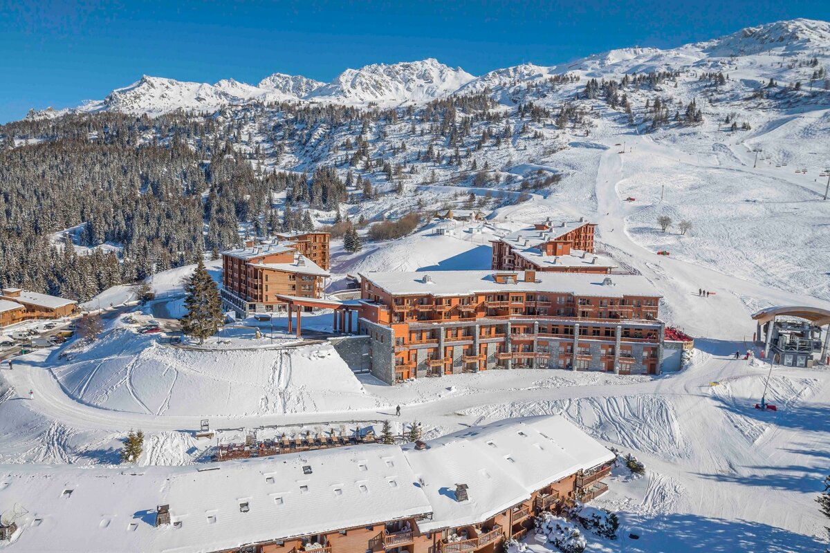 Aerial view of a snowy mountain ski resort with numerous wooden buildings, ski slopes, and pine forests under a clear blue sky.
