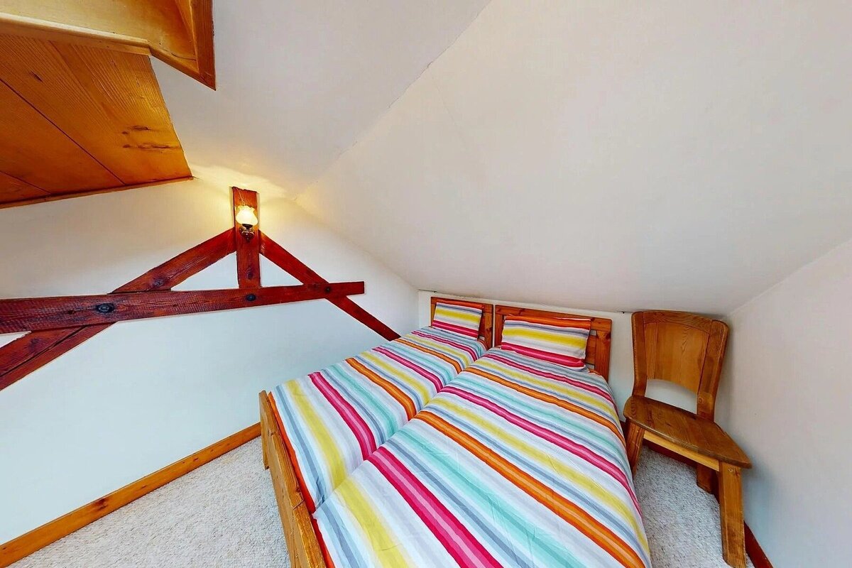 A cozy attic bedroom features two twin beds with colorful striped bedding, exposed wooden beams, a wooden chair, white walls, and carpet.